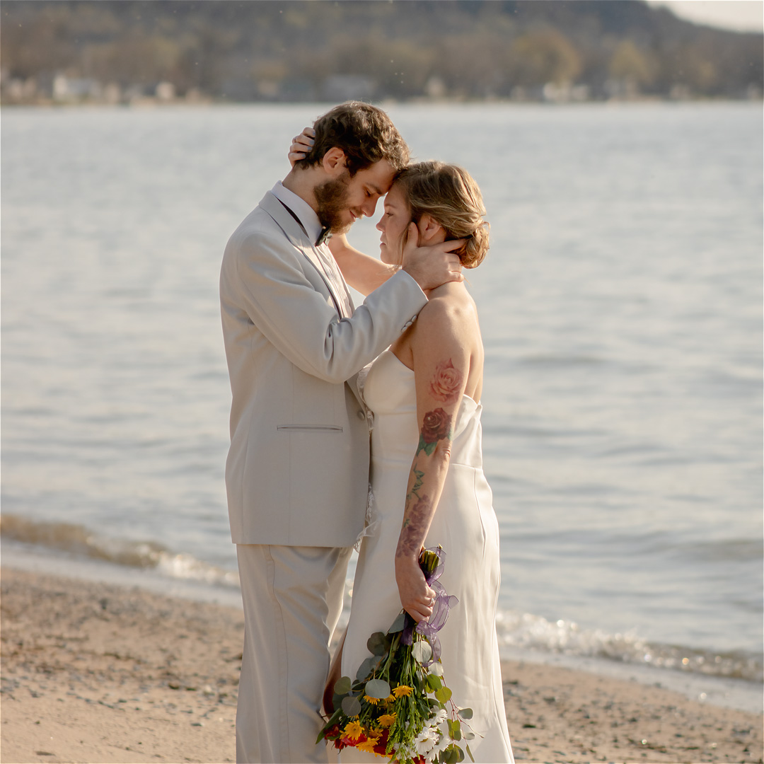 Bride and groom sharing a kiss on the beach. Bride holding bouquet. Watertown Wedding Photographer Westcott Beach Sackets Harbor New York CNY bridal photography North Country beach landscapes Portrait of a bride and groom. New England Weddings