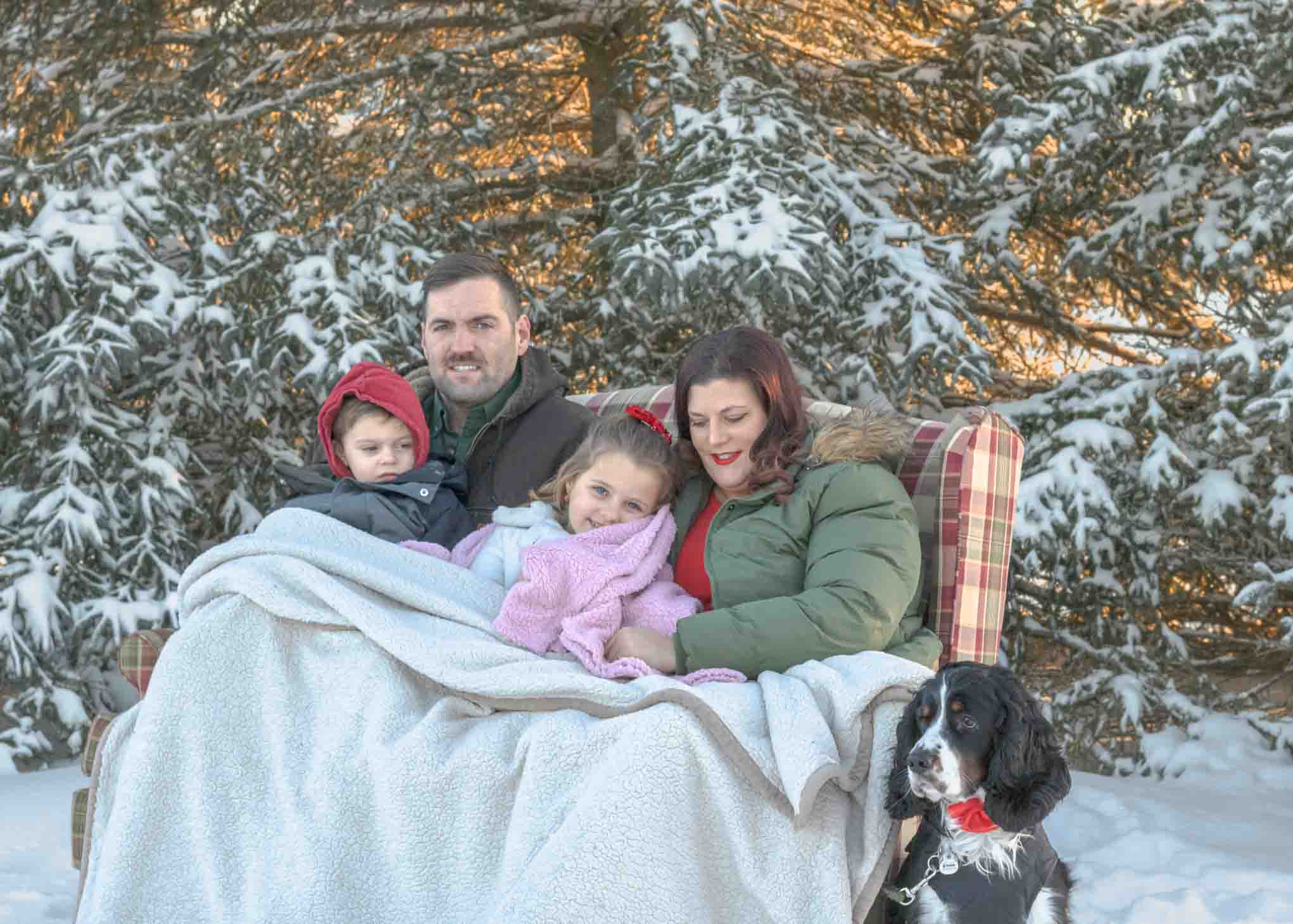 Family Portrait. Outdoor mom, dad, 2 kids and dog (spaniel) wearing a red bow-tie. Posed on couch with snow covered pine trees in the background. Military family photos Fort Drum Ny Northern New York CNY  Watertown NY