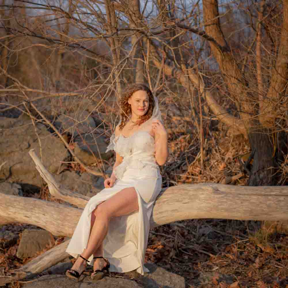 Bridal photography- editorial style bridal photo- portrait of a bride sitting on a fallen tree by the beach, white wedding gown, Lake Ontario. Golden hour- bright, light and airy photography. Westcott Beach, Alexandria Bay New York