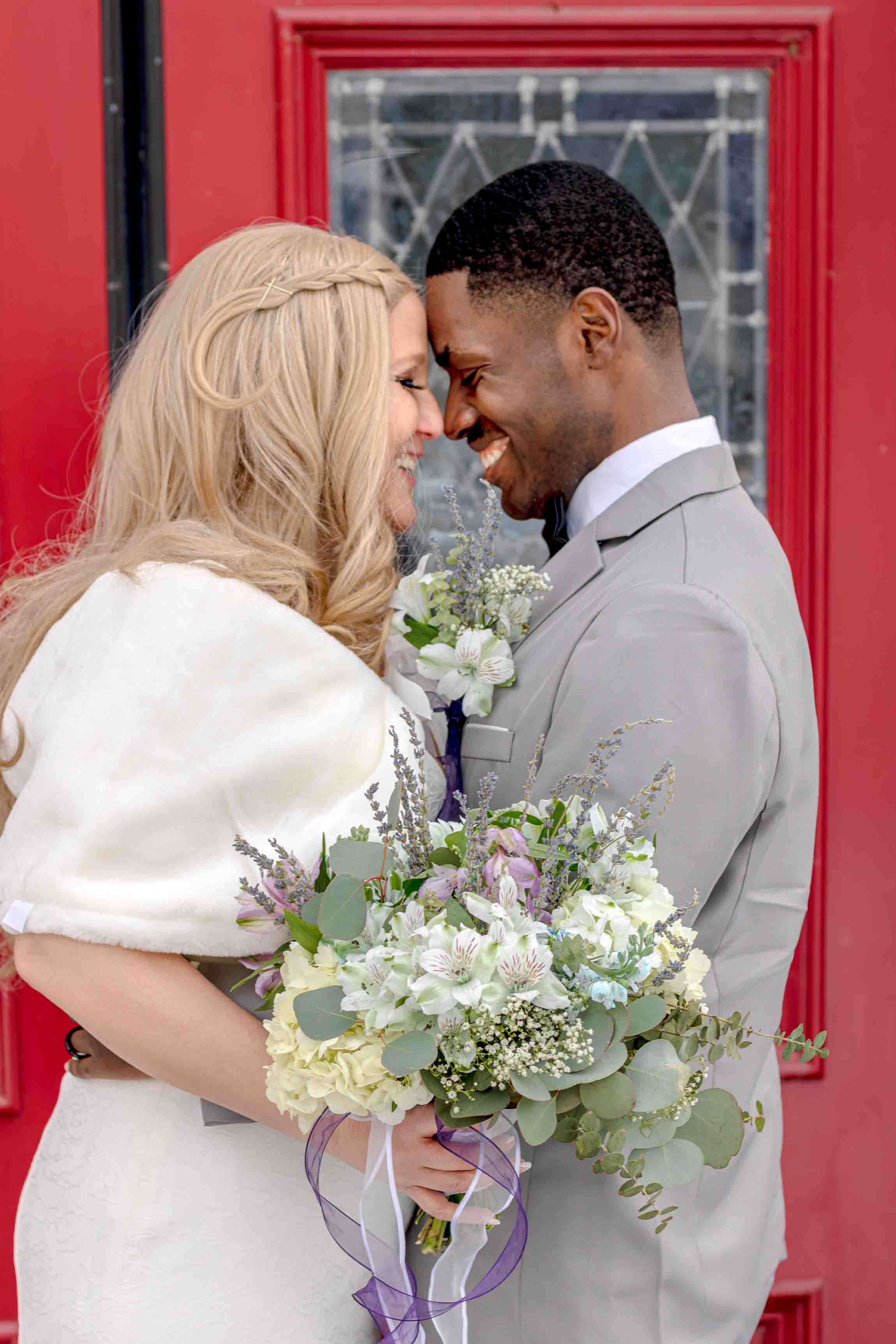 interracial couple, newlyweds, standing front of red church doors smiling at each other, bride holding bouquet Skaneateles, NY. CNY. Central New York. The Hamptons Long Island Brides. New England luxury Weddings. Northern New York Brides.