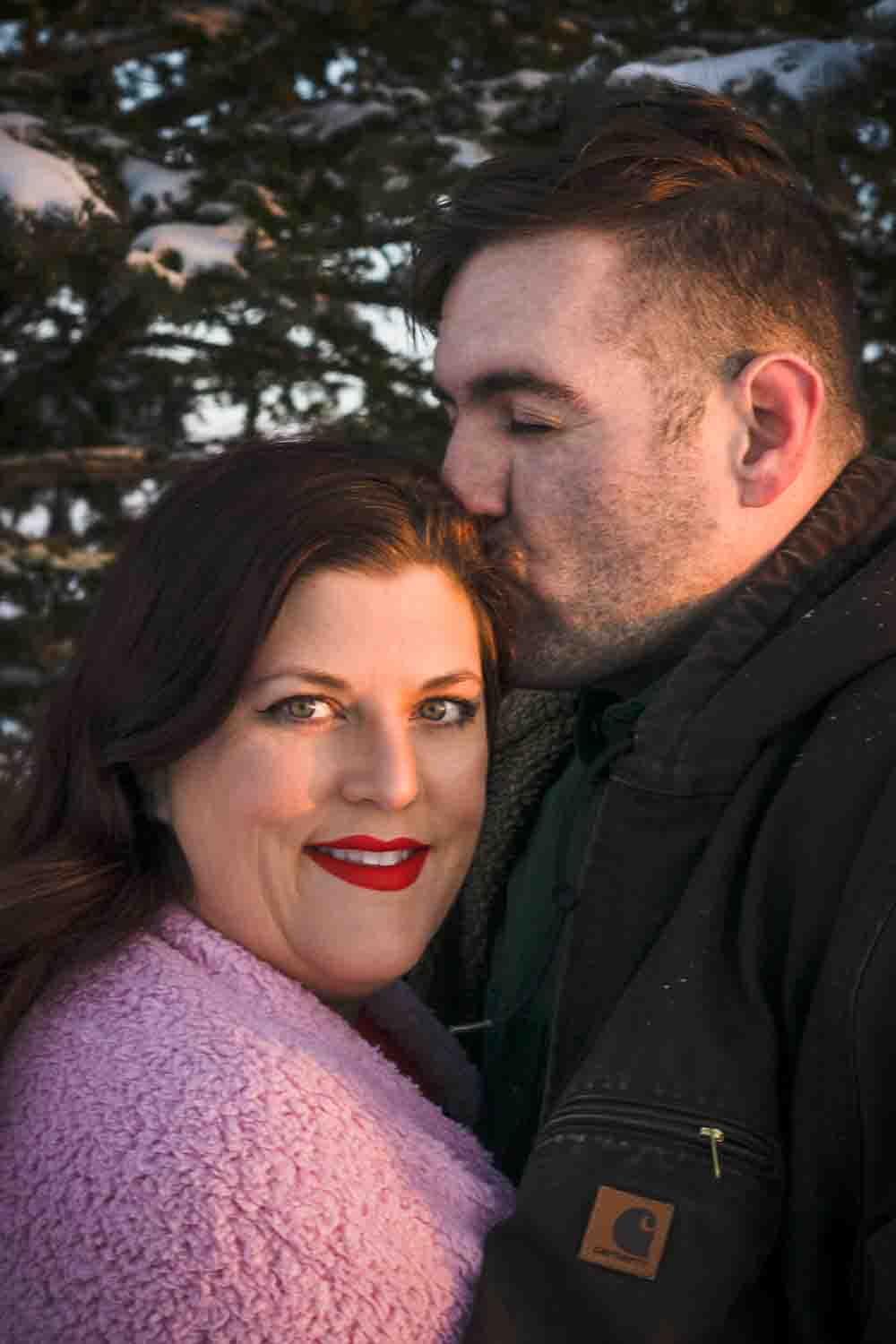 couples portrait photoshoot closeup headshot Loving couple man kissing woman's forehead with snow covered pines in the background Fort Drum Photographer