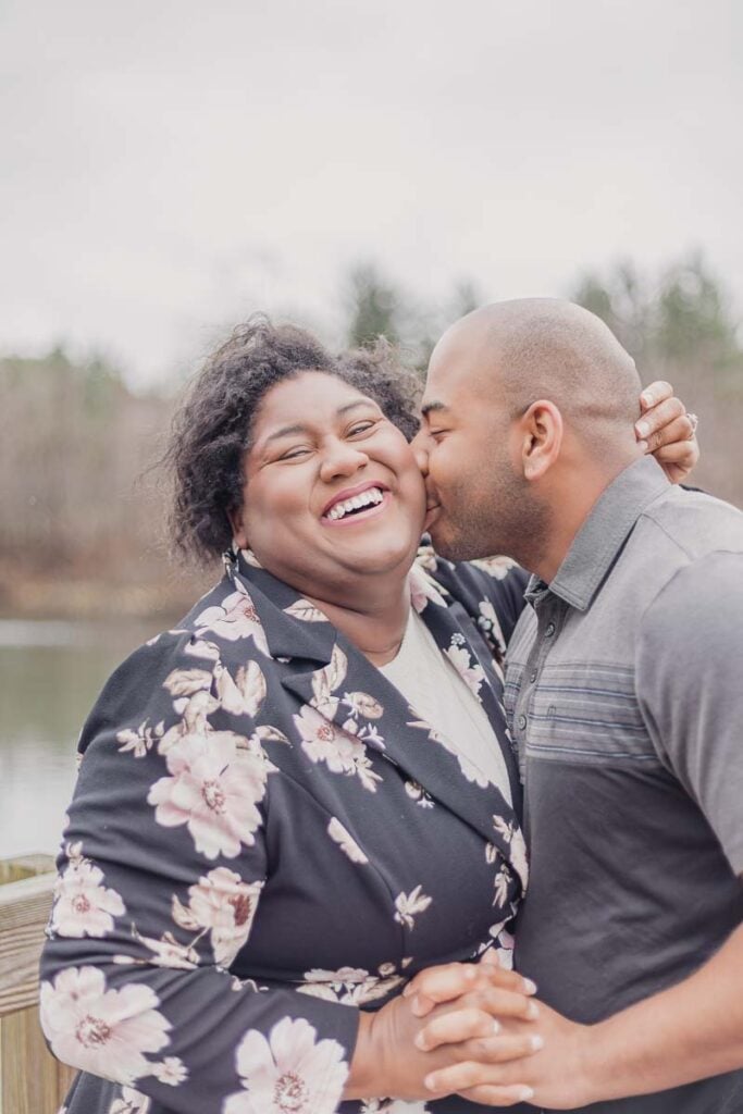 A joyful couple smiling as the man kisses the woman on the cheek while standing near a lakeside.