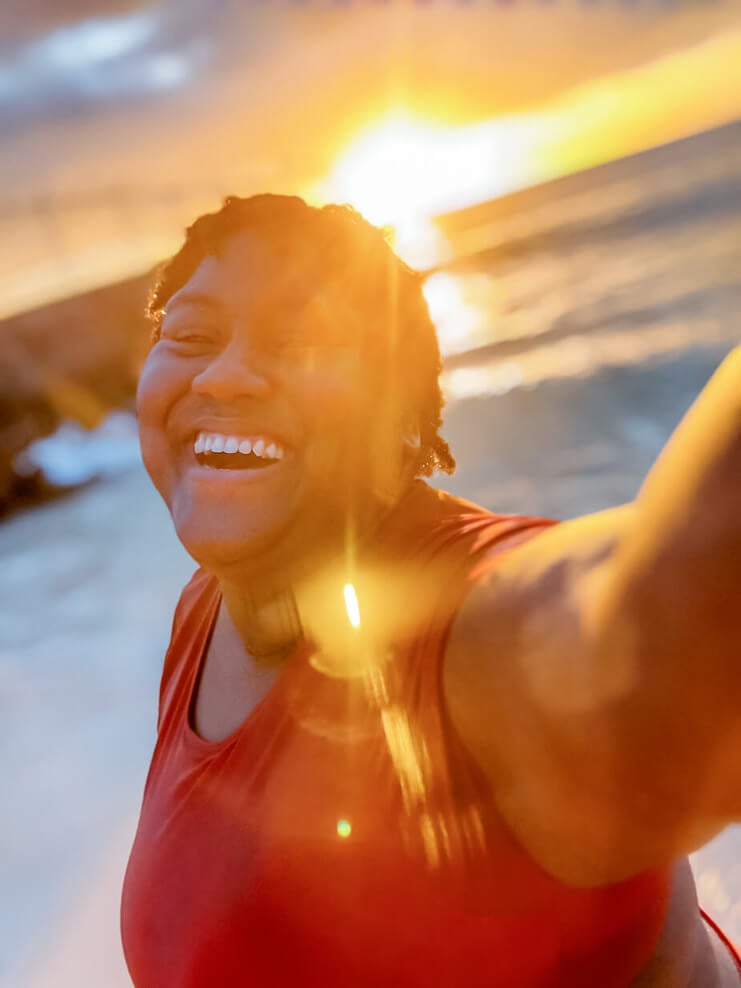 Traveling Wedding Photographer LaDonna Takyi Taylor smiling joyfully at the beach during sunset, golden sunlight glowing behind her.