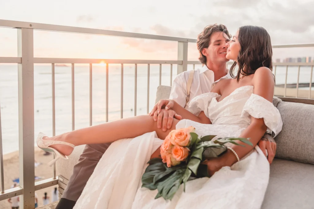 Bride and groom relax on an oceanview lounge at sunset with a peach rose bouquet at Kaimana Beach Hotel
