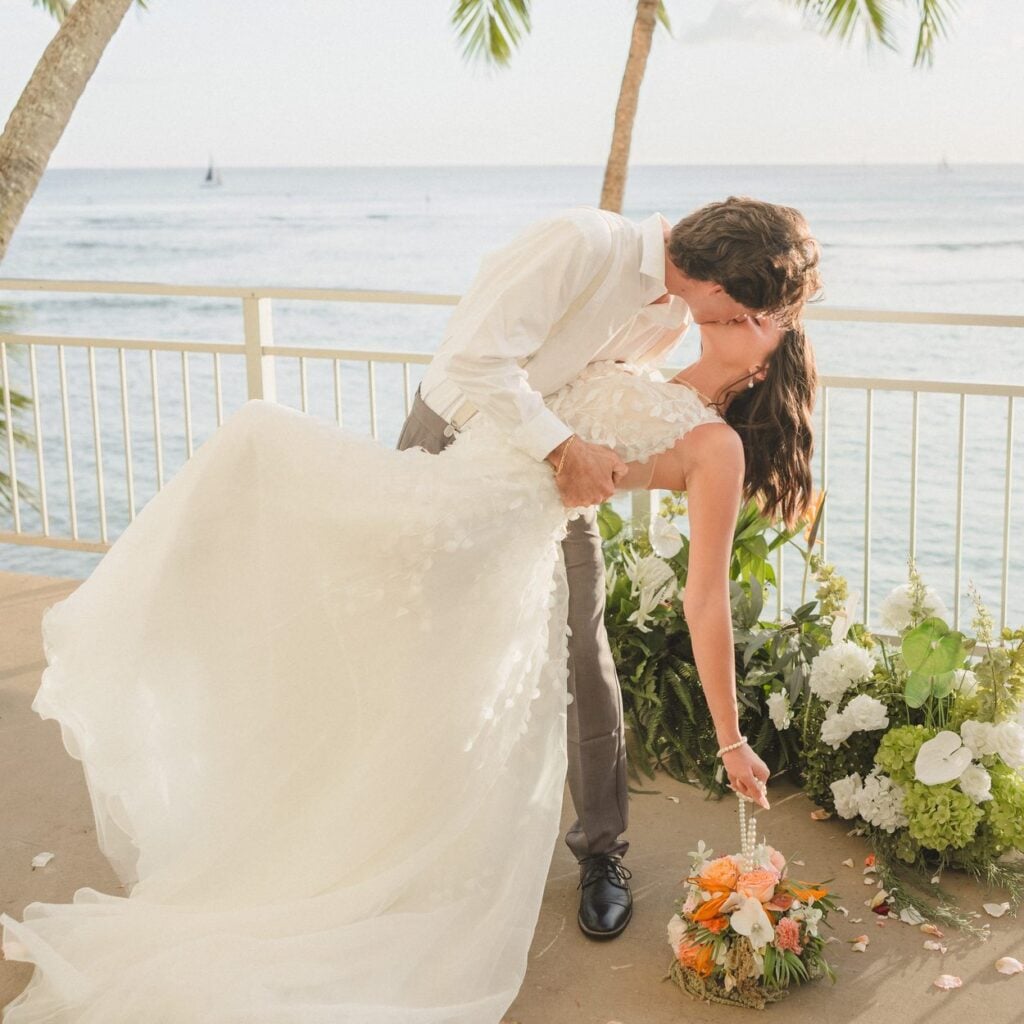 Groom dips bride for a kiss beside tropical ceremony florals and palm trees at an oceanfront Kaimana Beach Hotel wedding