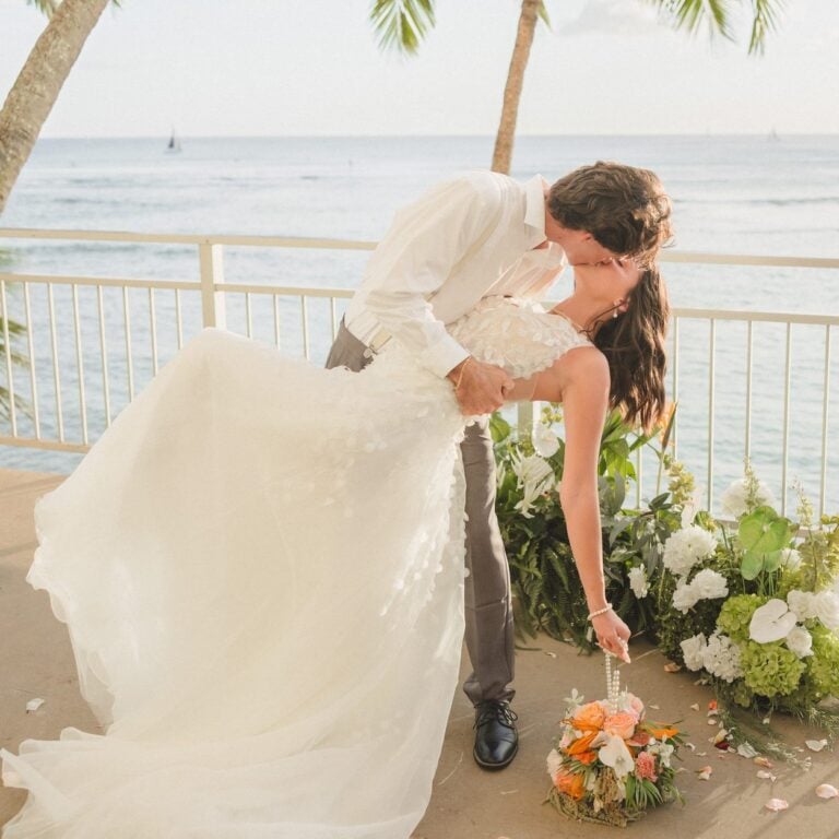 Groom dips bride for a kiss beside tropical ceremony florals and palm trees at an oceanfront Kaimana Beach Hotel wedding