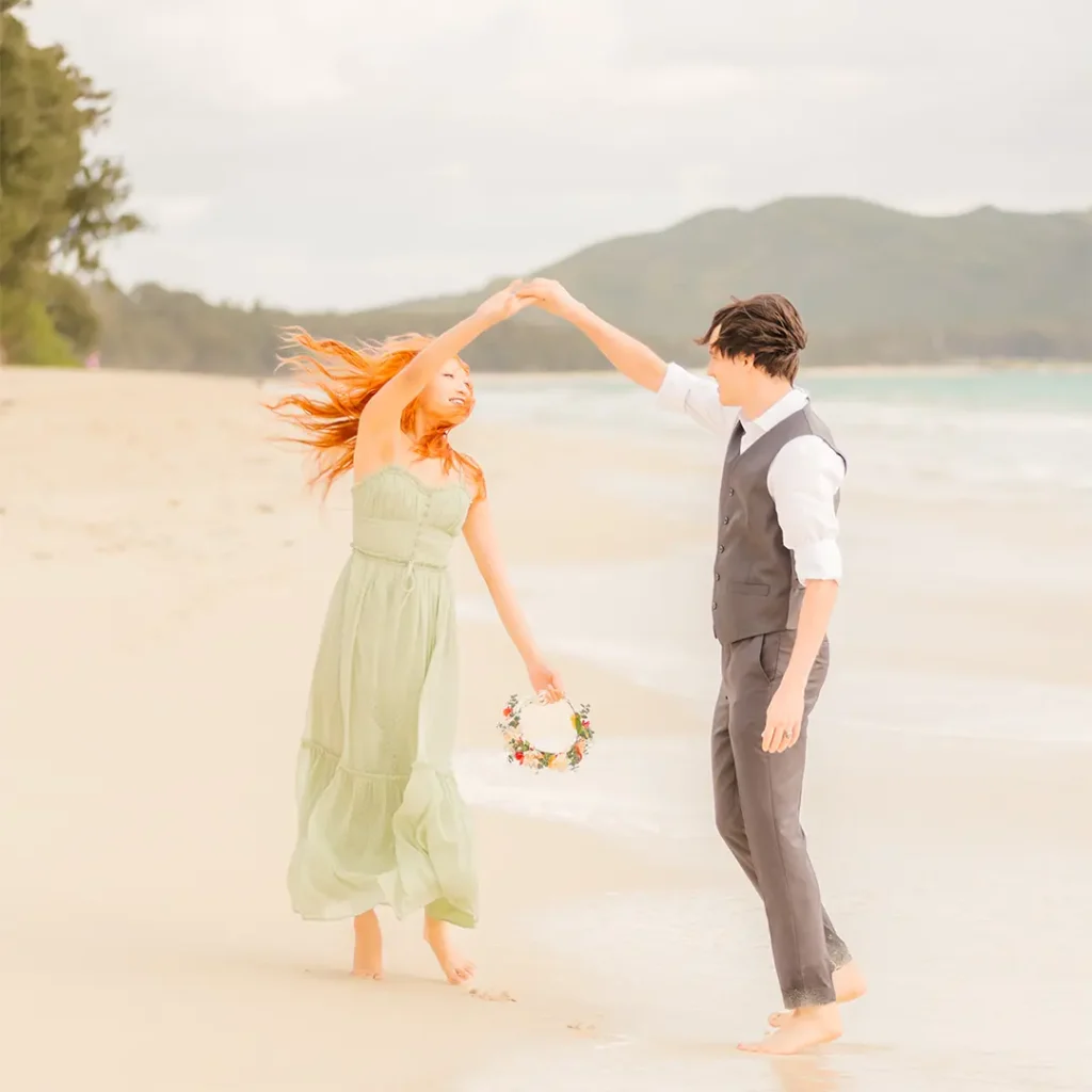 Couple dancing barefoot on the beach, holding hands and a floral crown during their Oahu engagement session.