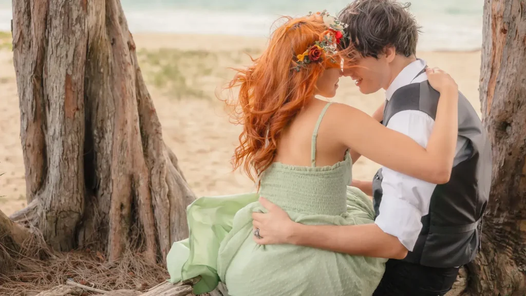 Engaged couple embracing beneath twisted tree trunks at a Hawaii beach.