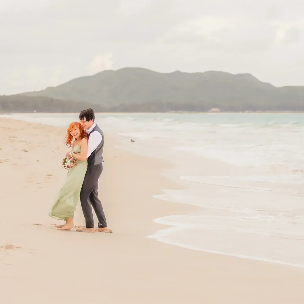 Couple holding each other and smiling as they walk barefoot along Waimanalo Beach.