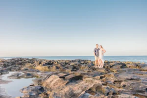 Traveling wedding photographer. Couple poses together on lava rocks with ocean and sunrise in the background