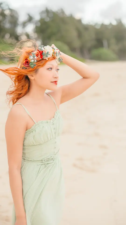 Oahu engagement photo of Senne standing barefoot on the beach in a sage green dress and floral crown, her red hair blowing in the wind.