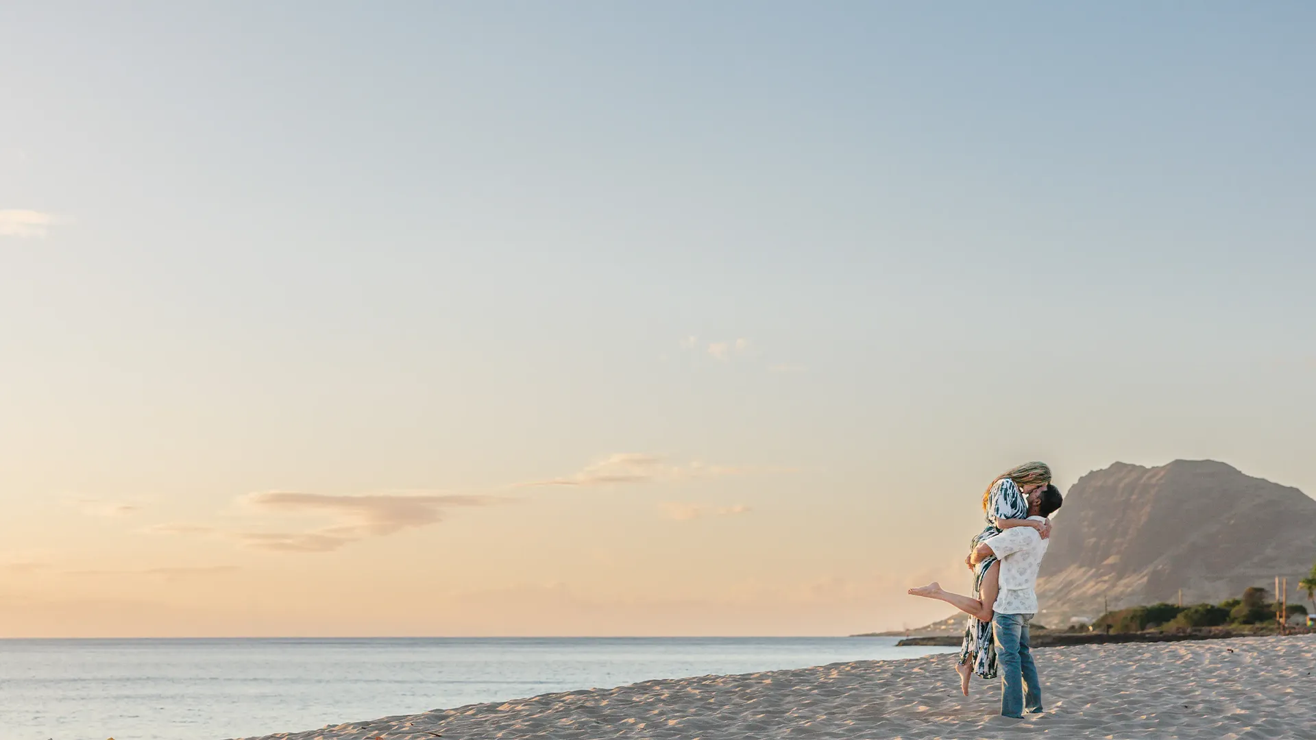 oahu proposal photographer. Man lifts fiancée on a beach at sunset after proposing during a Grand Gesture experience in Oahu