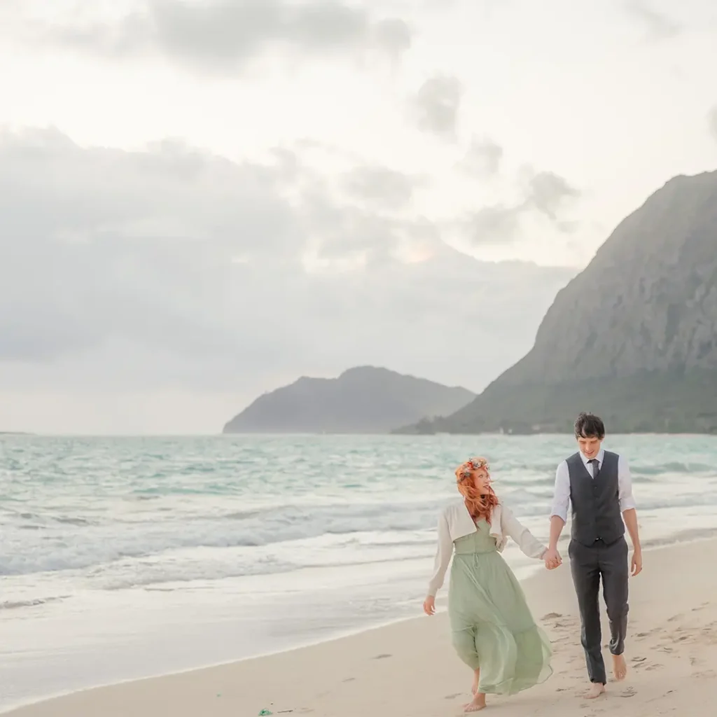 Engaged couple walking hand in hand along Waimanalo Beach, with ocean waves and distant mountains behind them.