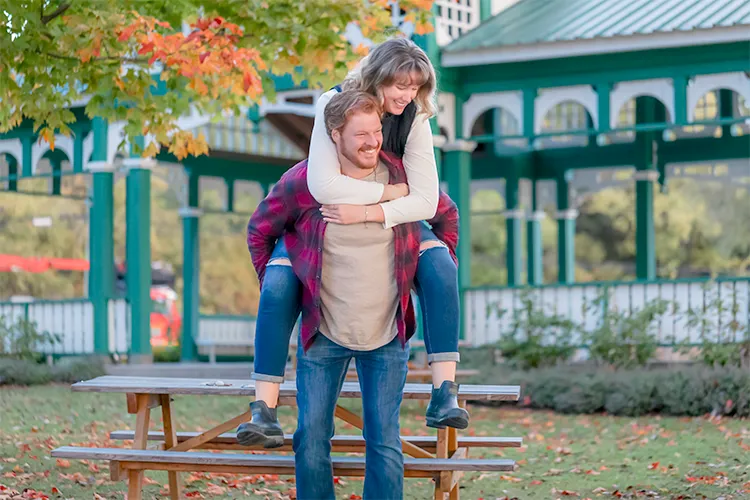 Fall engagement photoshoot. Dylan gives Hayley a piggyback ride beside a green gazebo during their fall engagement session.