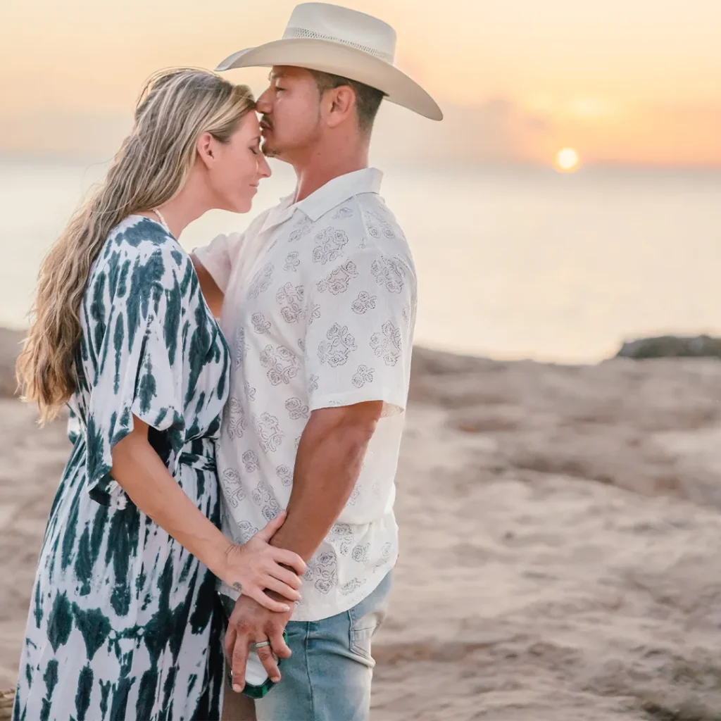 Cheyenne gently kisses Loraine’s forehead while holding her hand on a rocky beach at sunset in West Oʻahu.