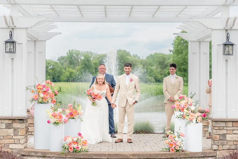 Summer weddings in Syracuse NY. Bride and groom holding hands under a white pergola during their outdoor wedding ceremony with vibrant floral arrangements and a fountain in the background.