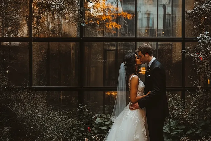 average cost of wedding photographer and videographer. Bride and groom share an intimate moment in front of a glass garden wall at a modern wedding venue.