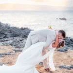 Groom dipping his bride for a kiss on a lava rock beach in Makena, Maui at sunset, her gown and sheer scarf trailing across the sand