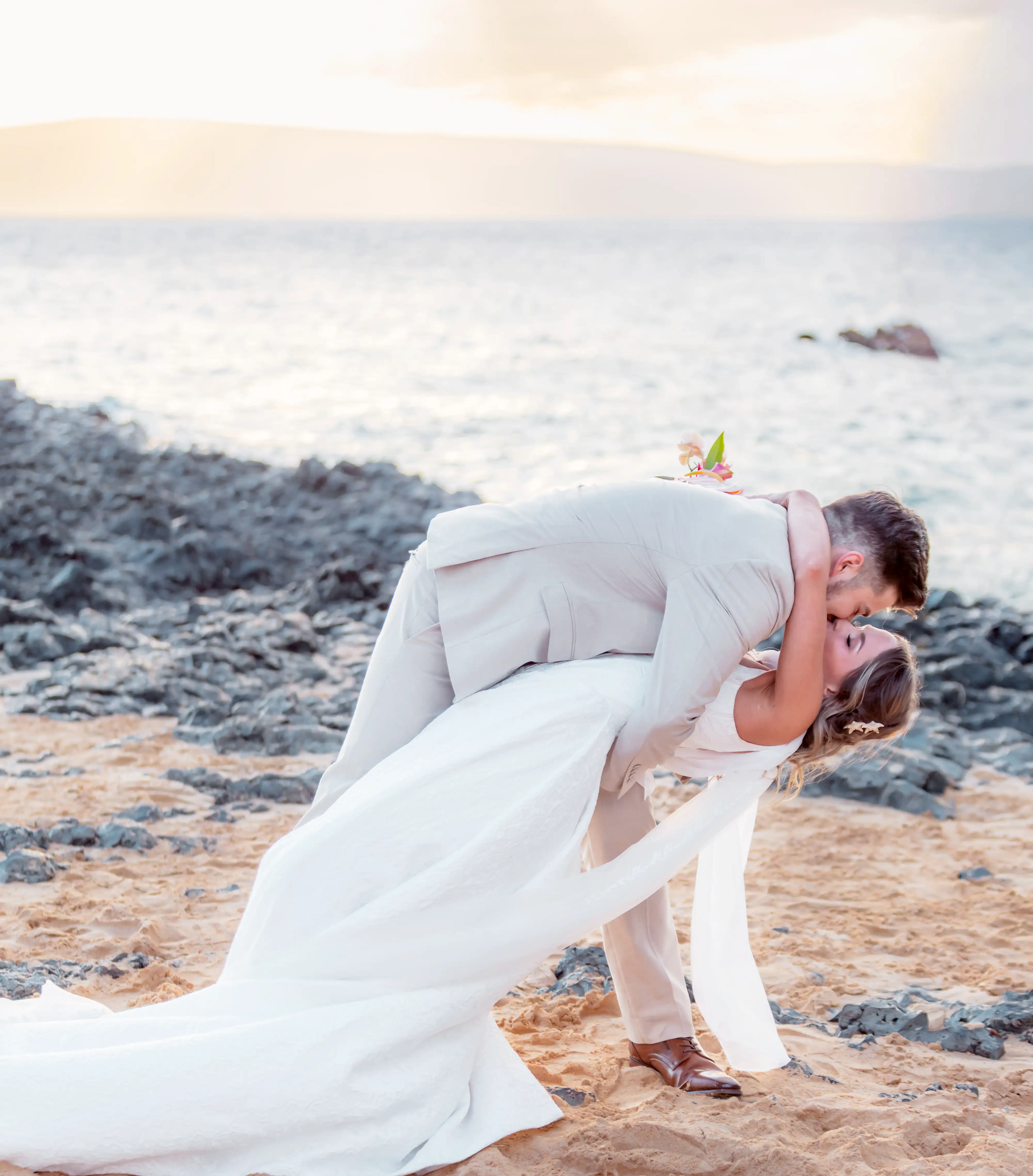 Groom dipping his bride for a kiss on a lava rock beach in Makena, Maui at sunset, her gown and sheer scarf trailing across the sand