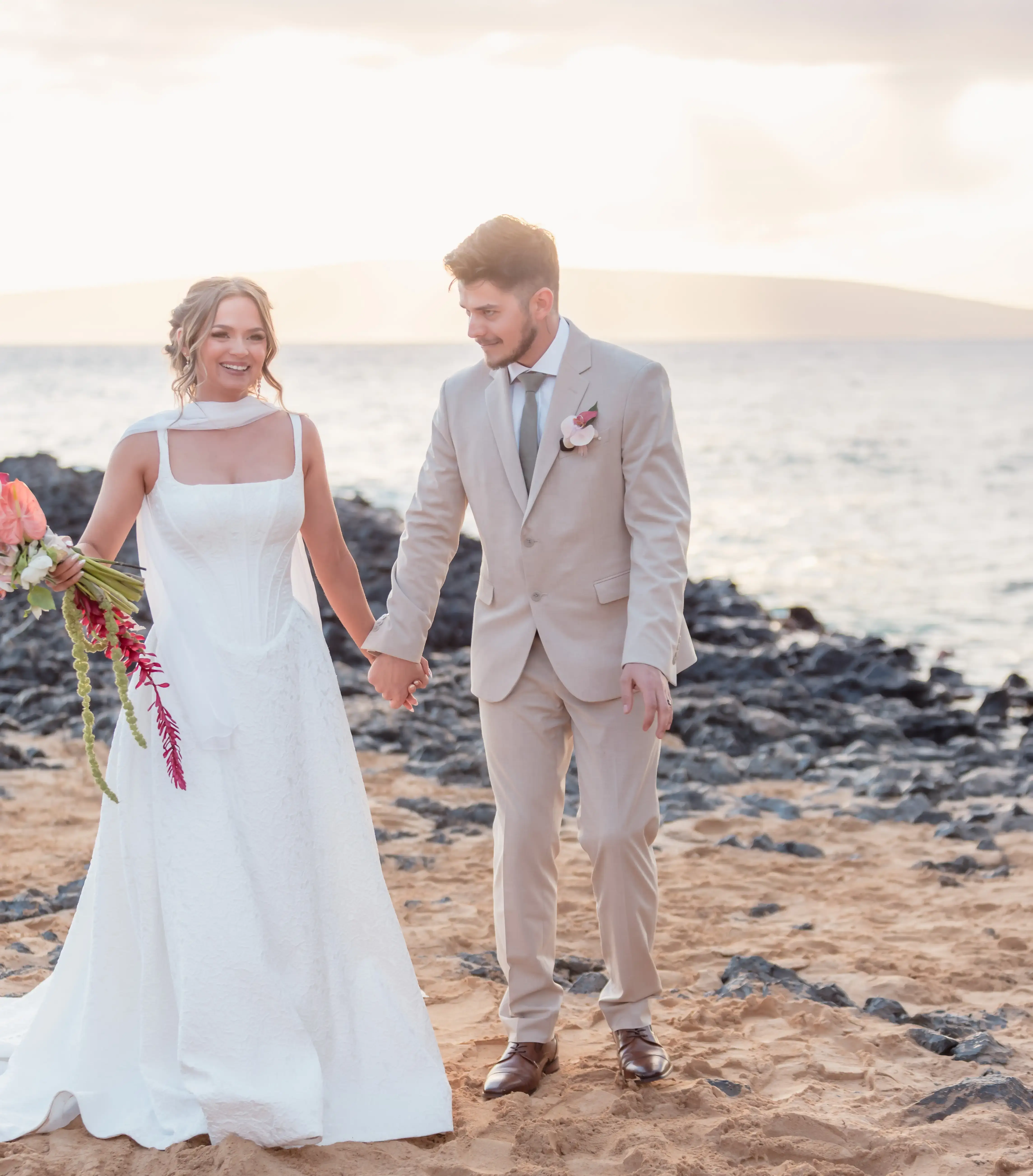 Bride and groom holding hands on a lava rock beach in Makena, Maui at golden hour, bride carrying a tropical bouquet