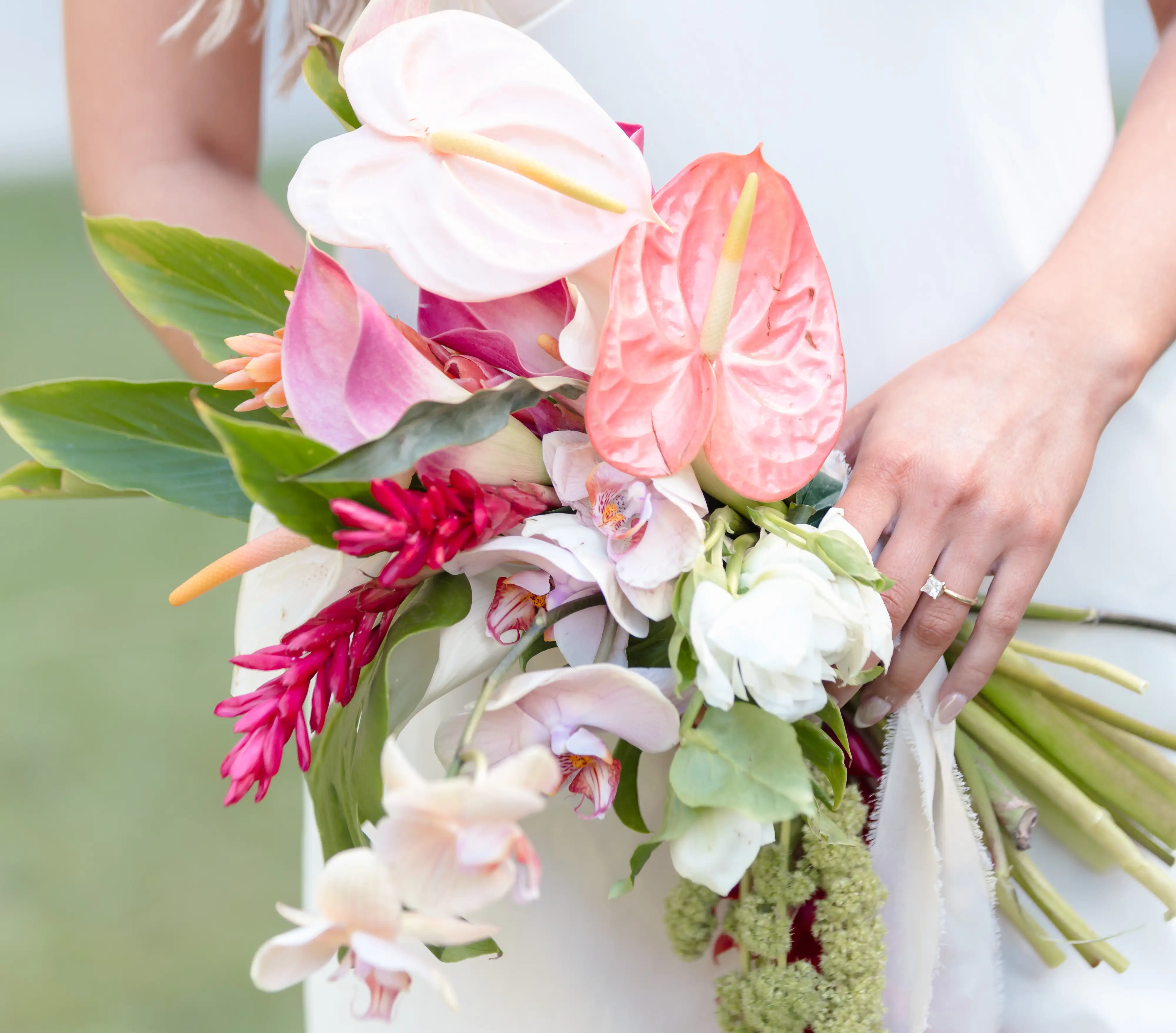 Bride holding a hand-tied tropical bouquet of blush anthurium, orchids, red ginger and green amaranthus against her white gown