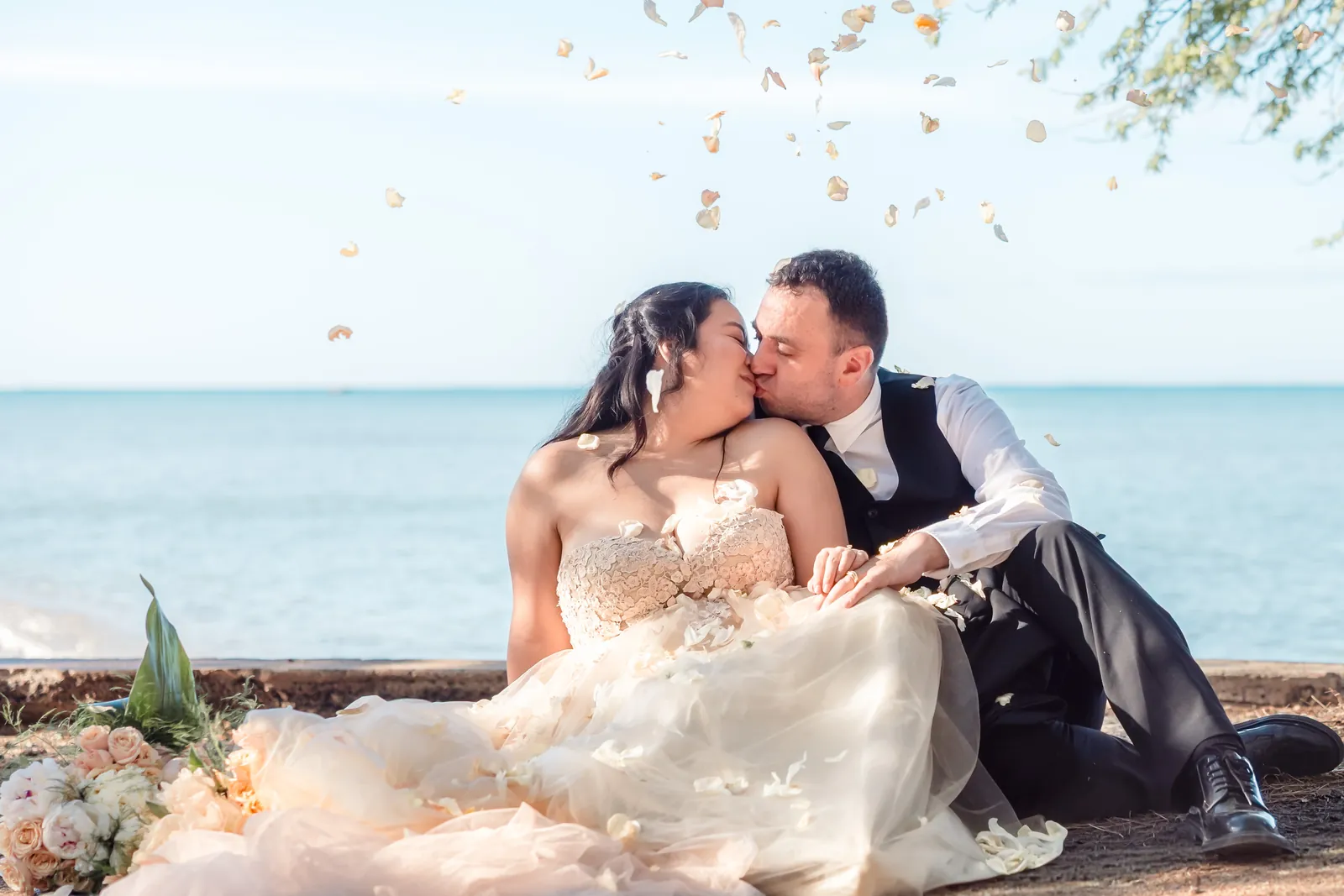 Oahu Hawaii Wedding Videographer, LT² Photography -Bride and groom kissing on the shore at Honeymoon Beach on Oahu as rose petals fall around them, her blush tulle gown spread across the sand