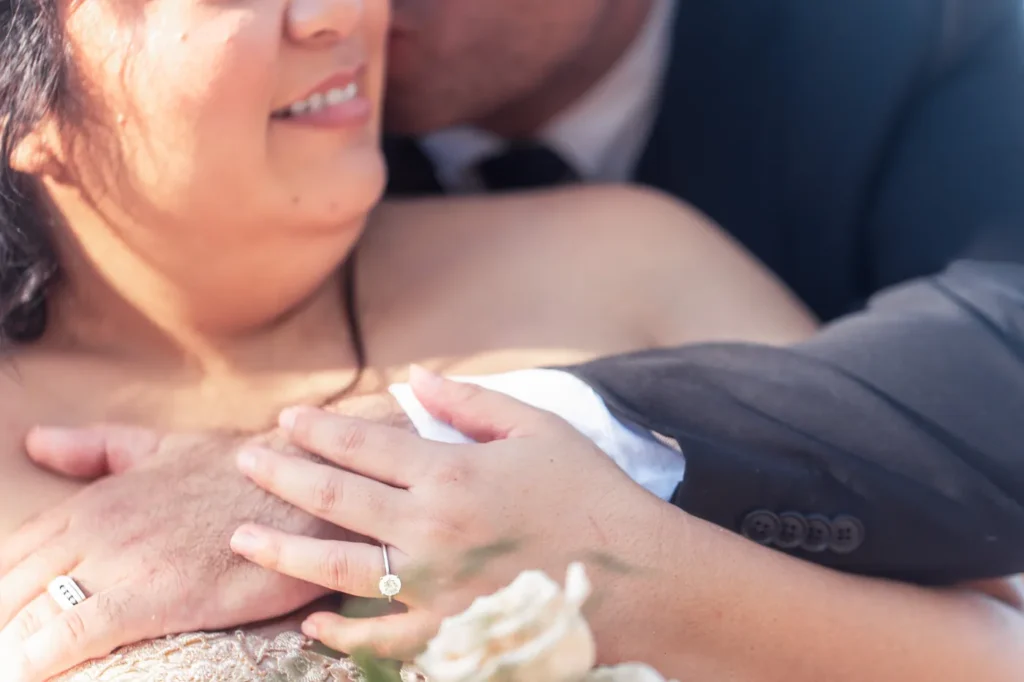 Close-up of the bride’s diamond ring as the groom kisses her cheek during wedding portraits at Hickam Beach on Oahu