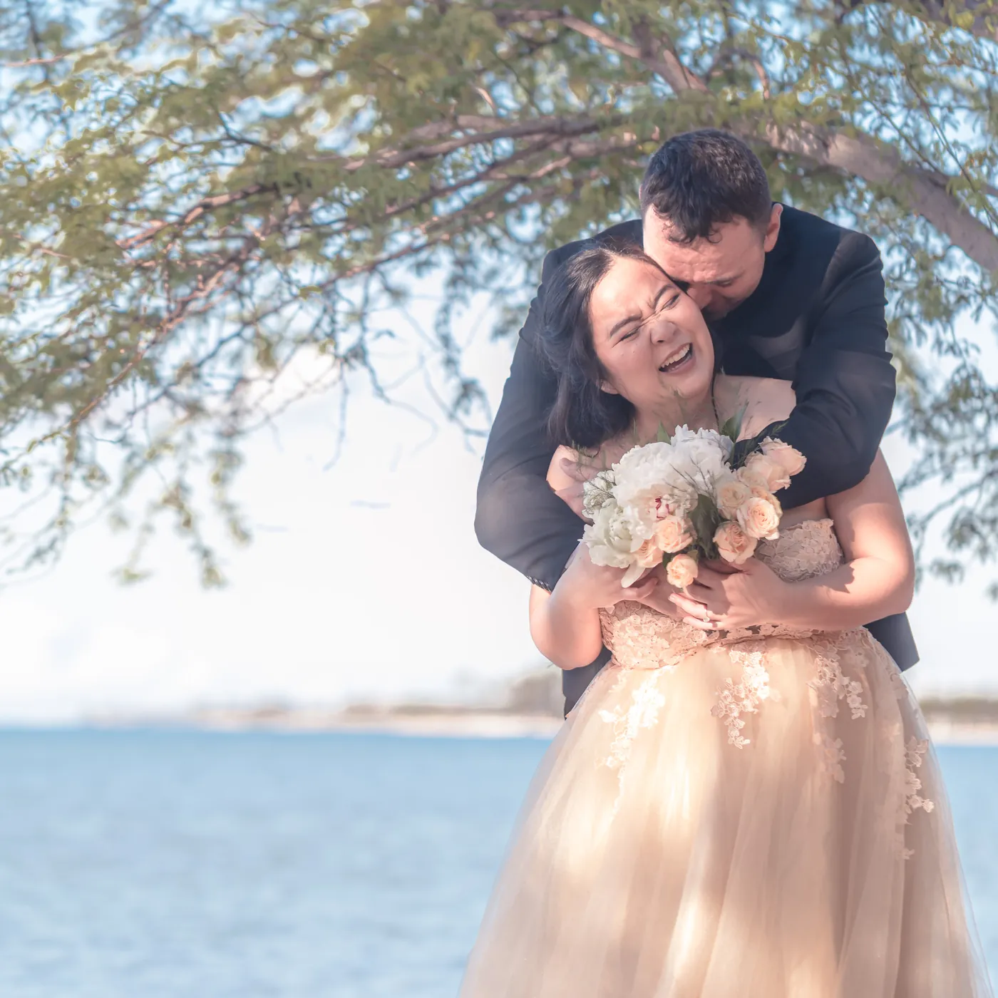 Bride laughing as the groom hugs her from behind under a shade tree at Hickam Beach on Oahu, holding a white and blush bouquet