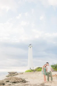 Engaged couple standing on the sand beneath the white Barbers Point Lighthouse under a soft cloudy sky in Kapolei, Oahu