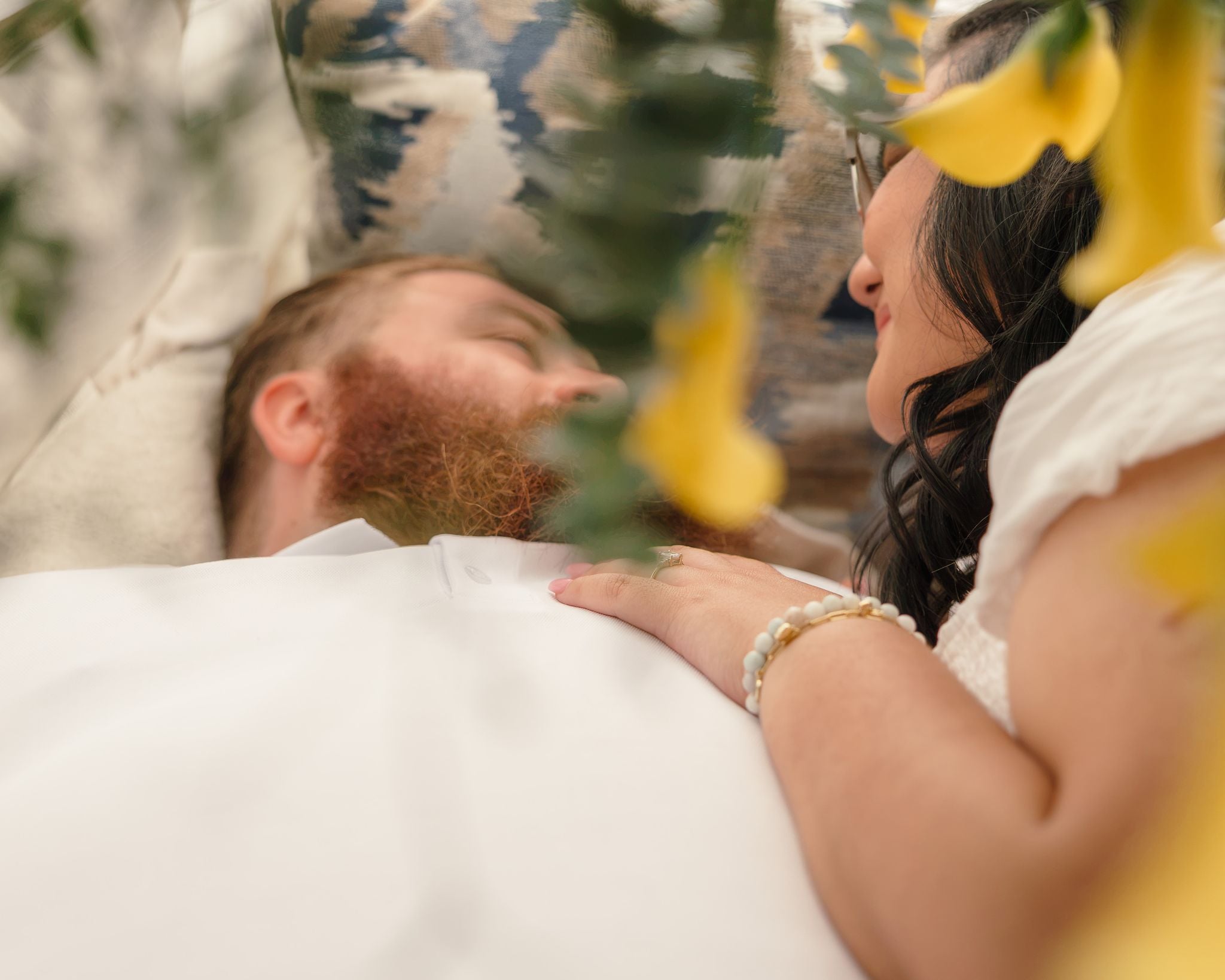 Dean lies back with his eyes closed as Hope rests her hand on his chest, framed by yellow calla lilies.