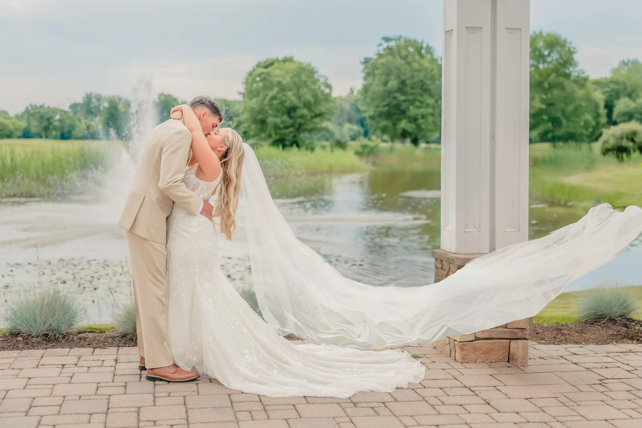 Bride and groom kissing beside a pond as her cathedral-length veil streams out sideways in the wind