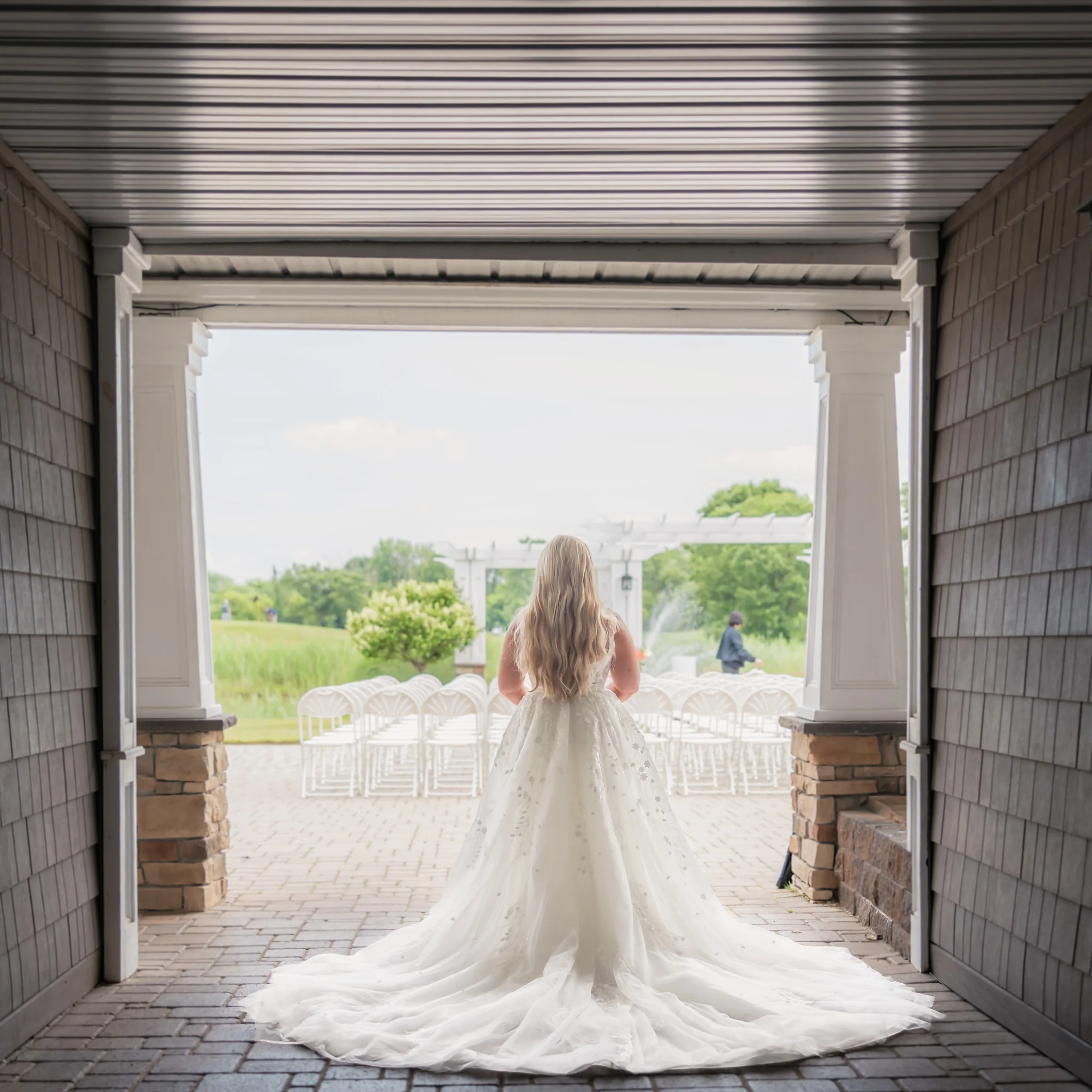 Bride seen from behind in a full ballgown, framed by a covered walkway as she looks out toward her outdoor ceremony