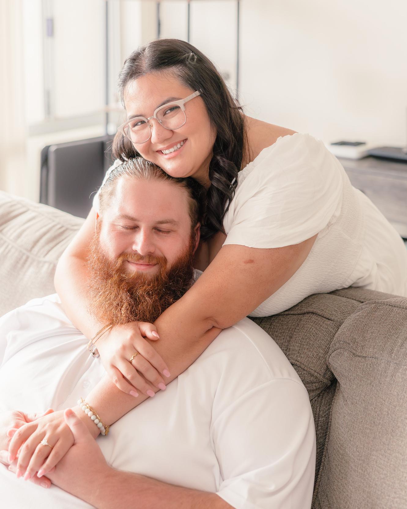 Hope hugs Dean from behind on the couch, her cheek on his head, smiling at the camera.