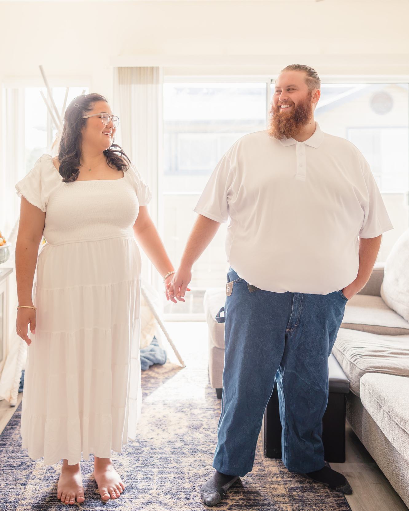 Hope and Dean hold hands and smile at each other in their living room, Hope barefoot on a patterned rug.