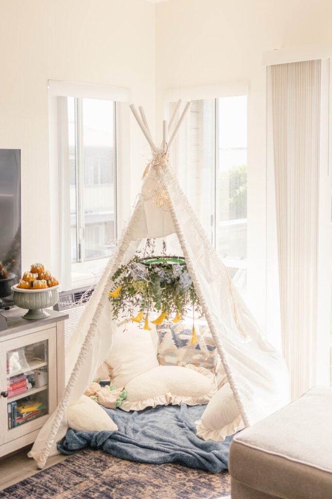 A white teepee with pillows, blankets and a hanging floral arrangement set up in a sunny living room for a date night photoshoot.