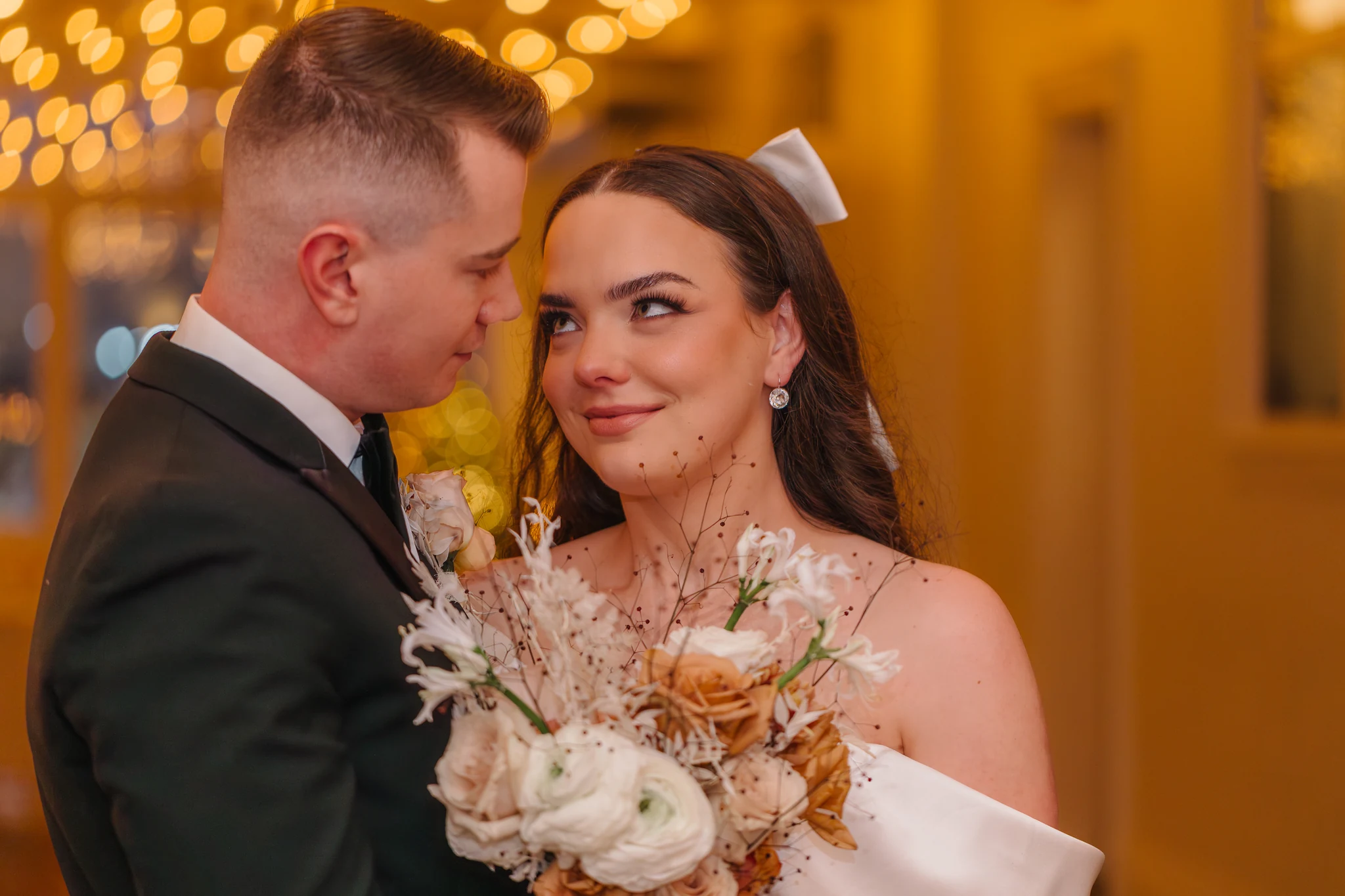 Groom leaning in toward his bride as she smiles at him over her bouquet, warm golden lights glowing behind them