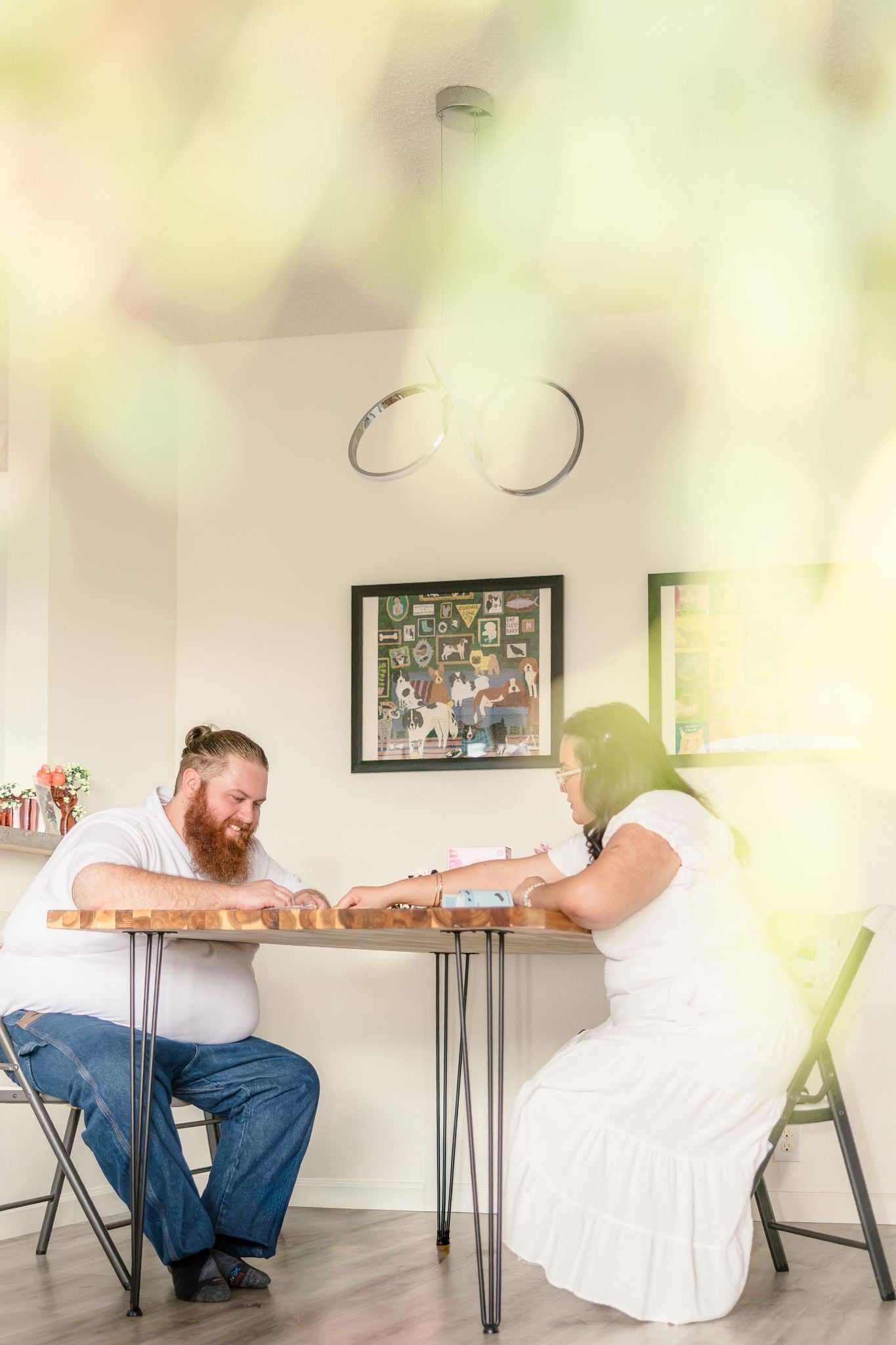 Dean smiles at Hope across the dining table during a game, seen through soft green leaves.