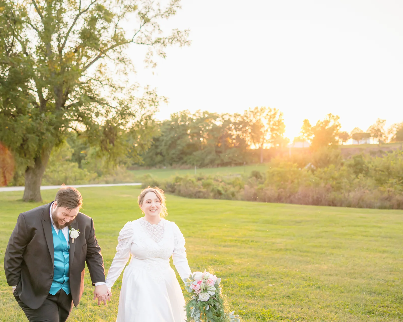 Bride and groom laughing as they walk hand in hand across a green lawn with the sun setting behind them