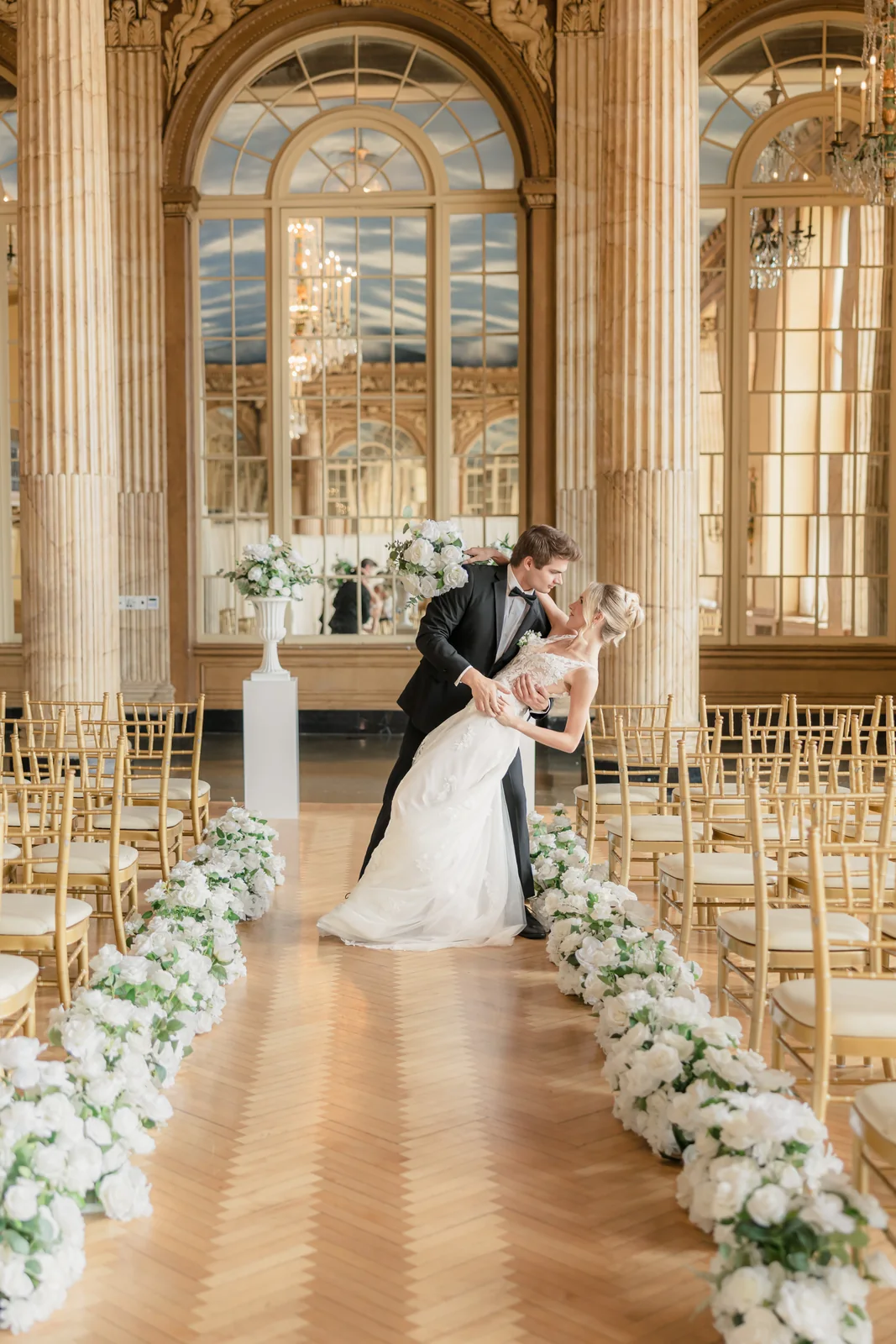 Groom dipping his bride into a kiss at the end of a white-flower-lined aisle in the gilded Hotel Syracuse ballroom