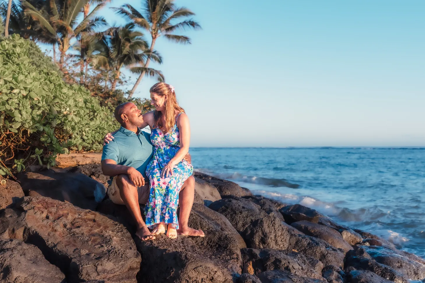 Engaged couple sitting together on the shoreline rocks at Kaʻaʻawa Beach with the ocean and palms behind them