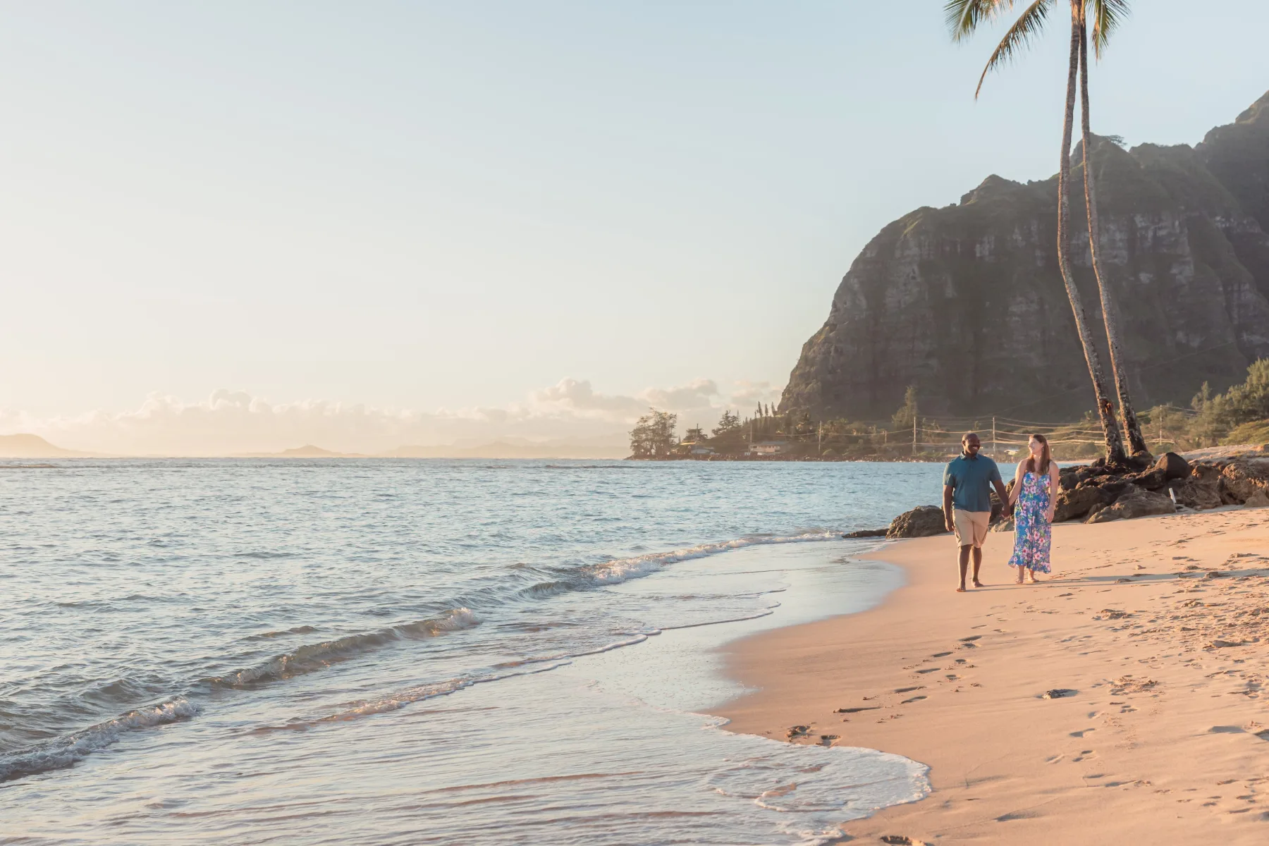 Engaged couple walking along Kaʻaʻawa Beach at sunrise with a palm tree and the Koʻolau mountains behind them, Oahu