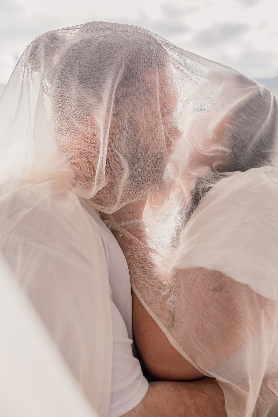 Hope and Dean kiss under a sheer canopy of fabric in soft window light.