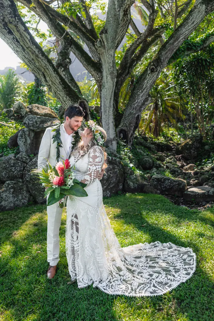 Bride in a plumeria haku lei and groom in a green lei beneath a large tree at Paliku Gardens, Kualoa Ranch, Oahu, her lace train across the lawn