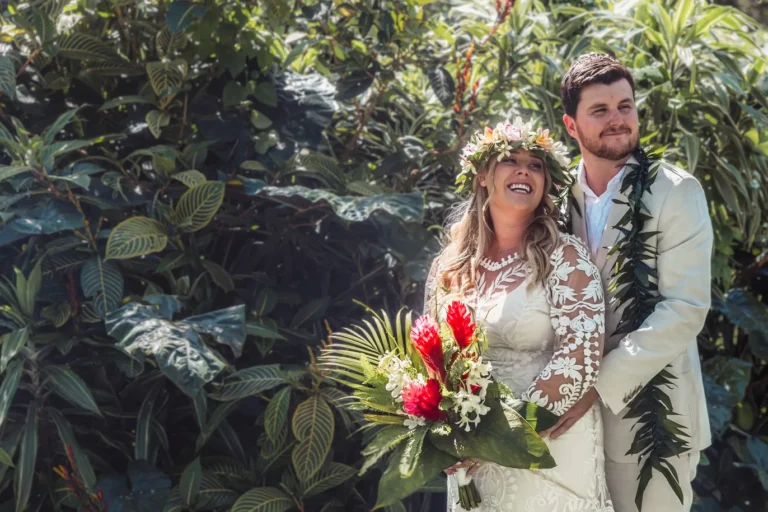 Bride laughing in a flower haku lei and lace gown, holding a red ginger bouquet, as the groom in a linen suit and green lei stands behind her among tropical foliage at Kualoa Ranch on Oahu