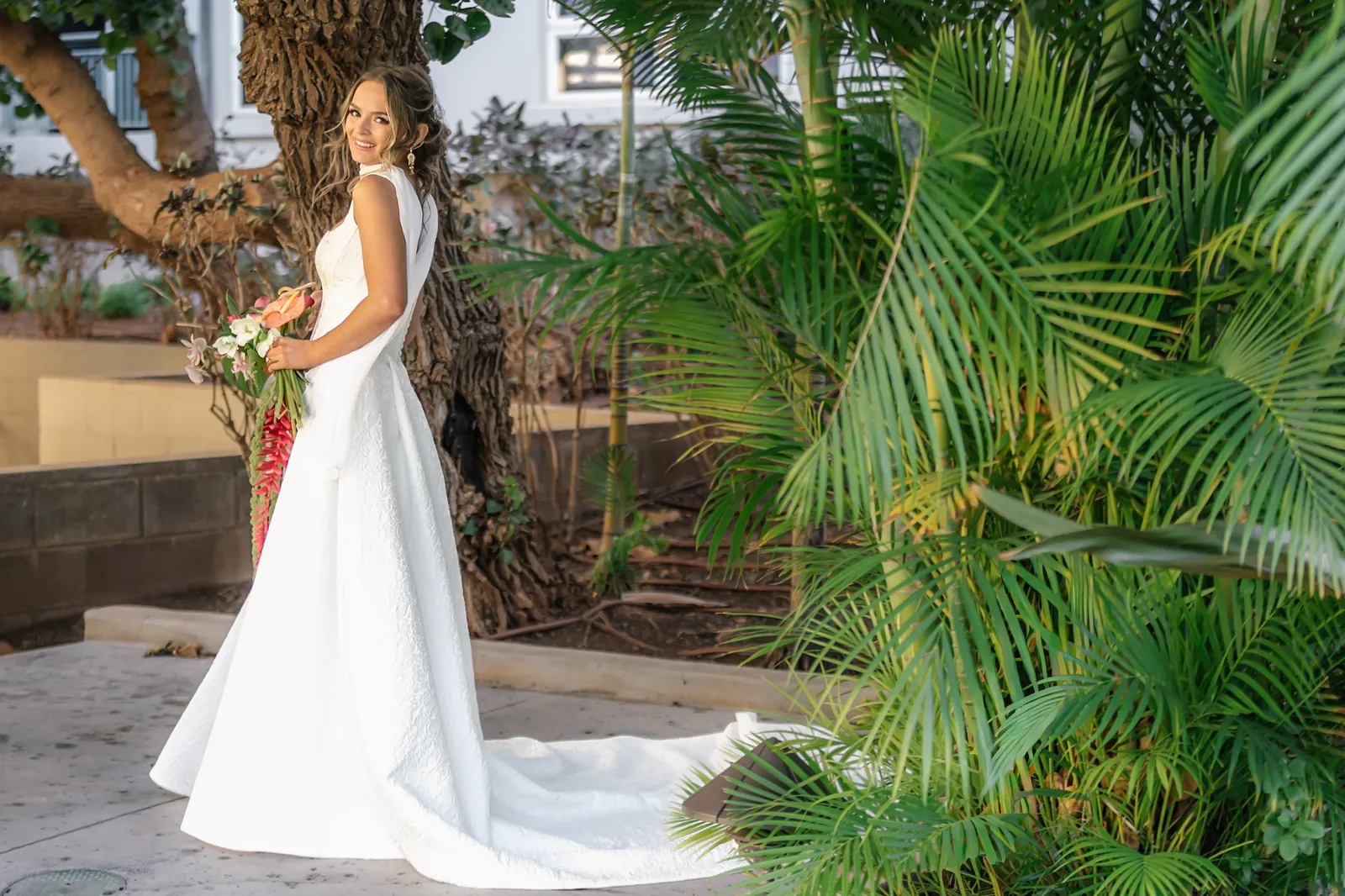 Bride in a sleeveless ivory gown with a long train holding a tropical bouquet among palms in a Maui resort garden on her elopement morning