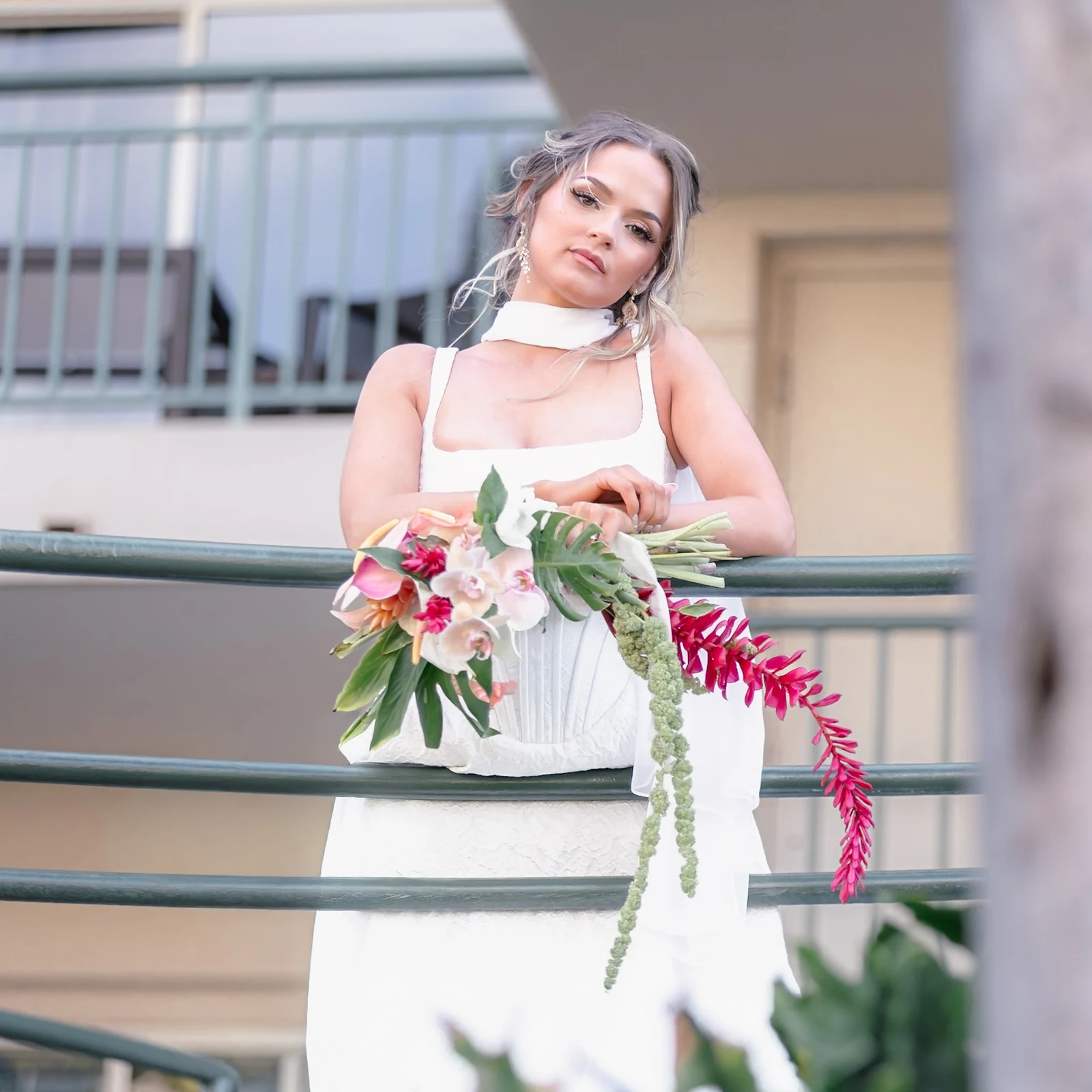 Bride leaning on a balcony railing holding a tropical bouquet of orchids, red ginger and hanging amaranth before her Maui elopement