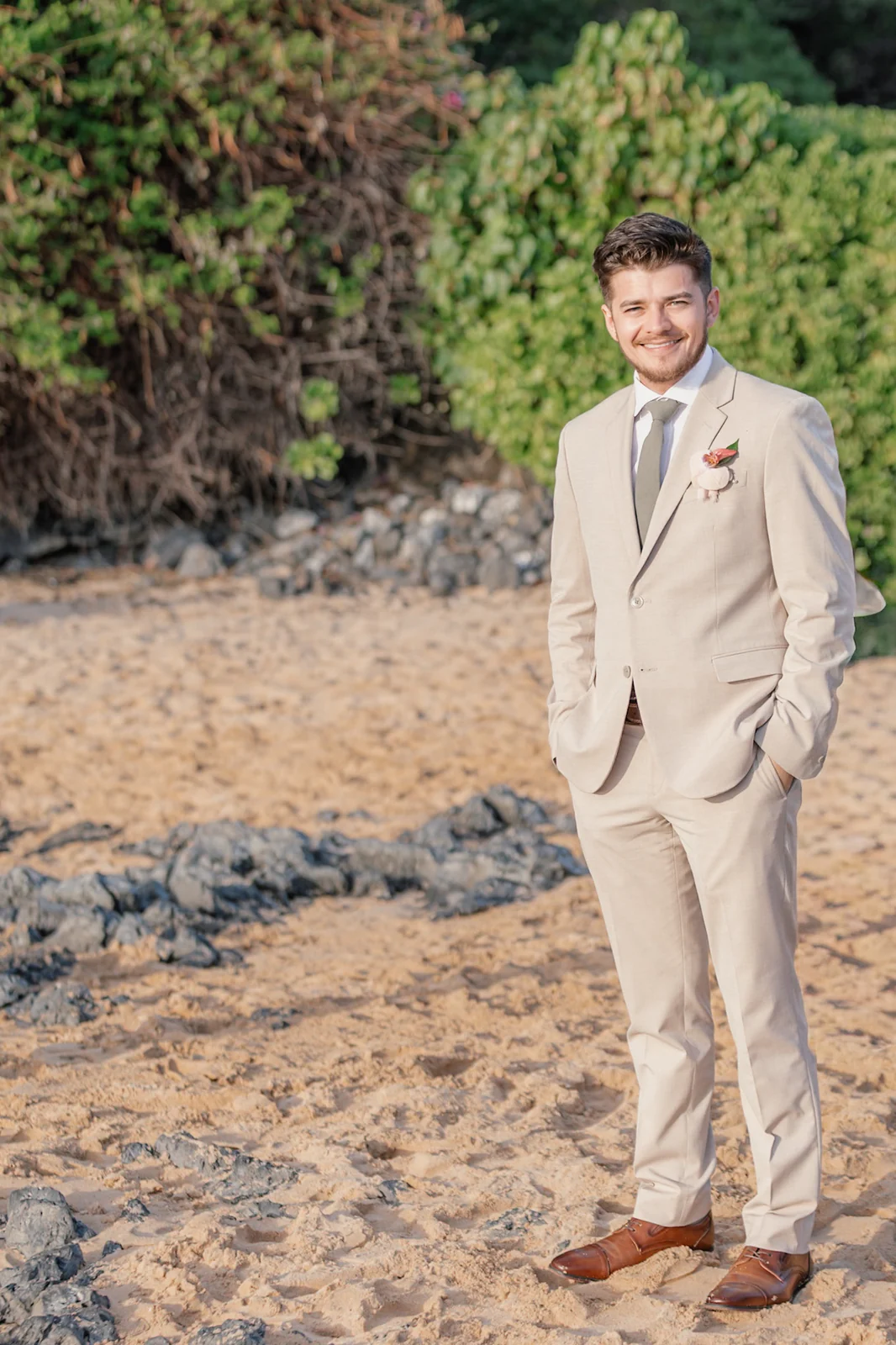 Smiling groom in a tan suit with hands in his pockets on the sand at Secret Cove beach in Makena, Maui
