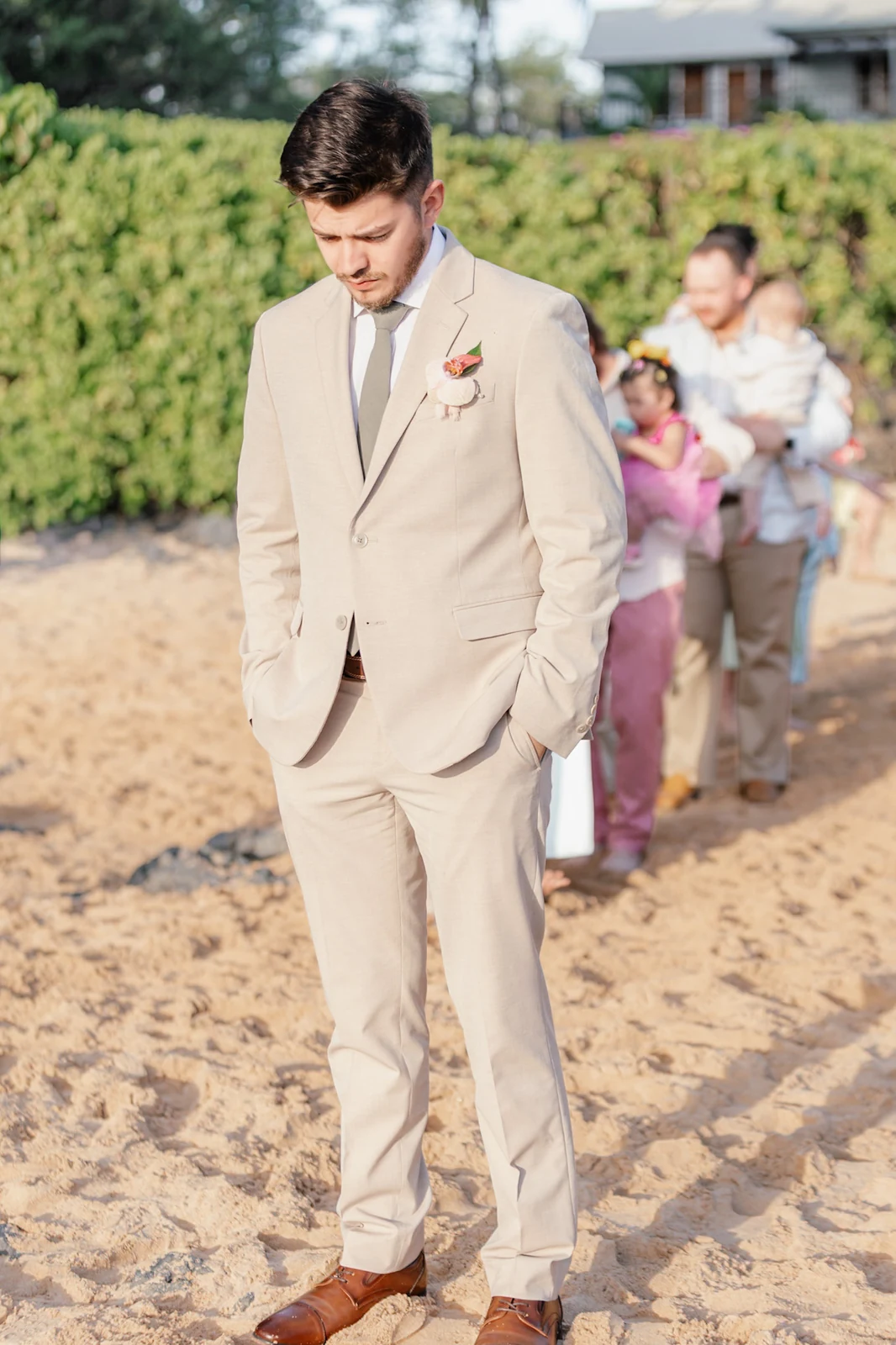 Groom in a tan suit waiting on the sand at Secret Cove in Makena as family gathers behind him before a Maui elopement ceremony