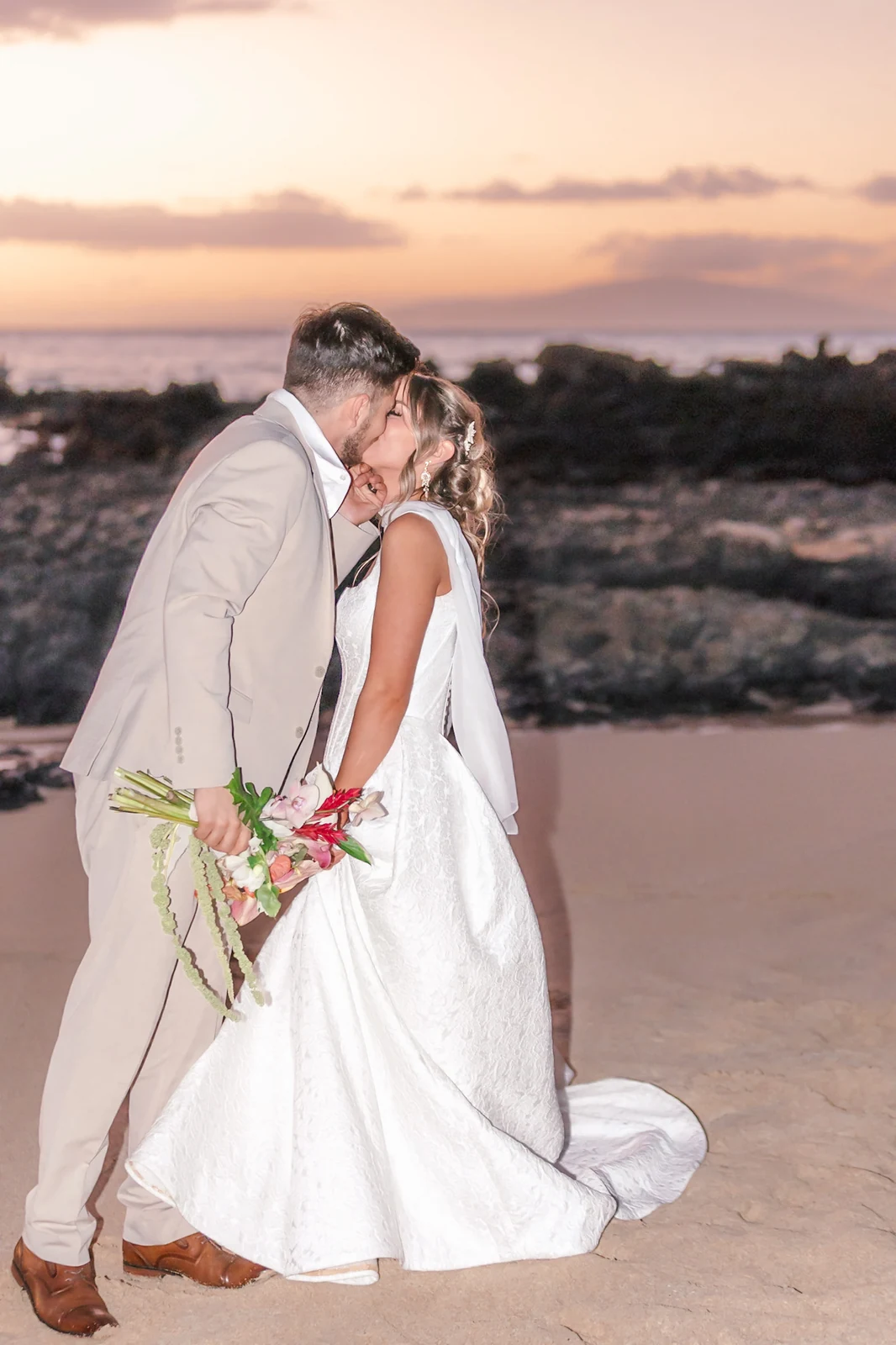 Newlyweds kissing on the beach at Secret Cove in Makena under a pink and orange Maui sunset with Kahoolawe on the horizon