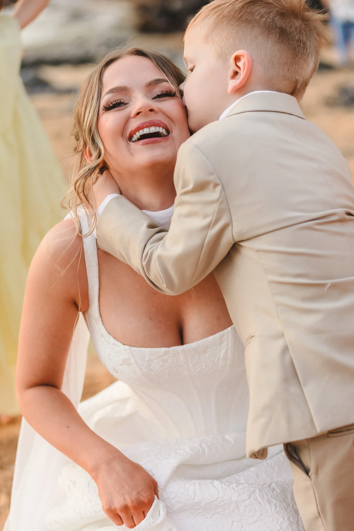 Bride laughing as a little boy in a tan suit hugs her and kisses her cheek on the beach